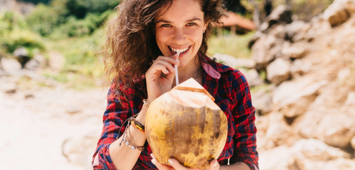 Happy enjoyable woman with adorable smile drinks coconut cocktail and smiles at camera on the sandy beach. Vacation mood with lovely young woman on the island Coconut water